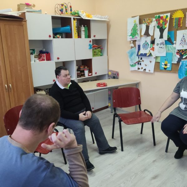 Trzej ludzie siedzący w okręgu, rozmawiają. W tle kolorowe prace dziecięce i szafki.