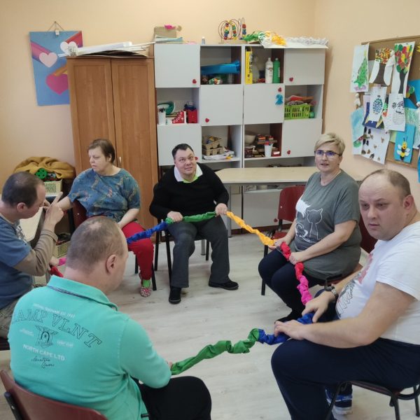 Grupa osób z niepełnosprawnościami siedzących w okręgu, trzymających wstążki.