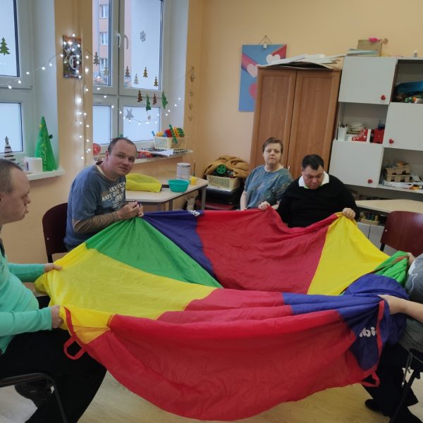 Cztery osoby trzymają kolorową tkaninę siedząc wokół niej, piąta osoba stoi obok.